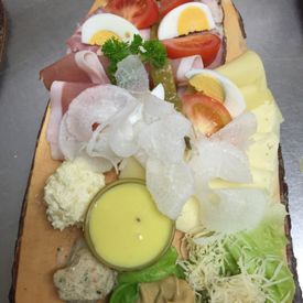 längliche Servierplatte aus Holz auf der Schinken, Käse, verschiedene Aufstriche, gekochte Eier und Salatbeilagen zu sehen sind
