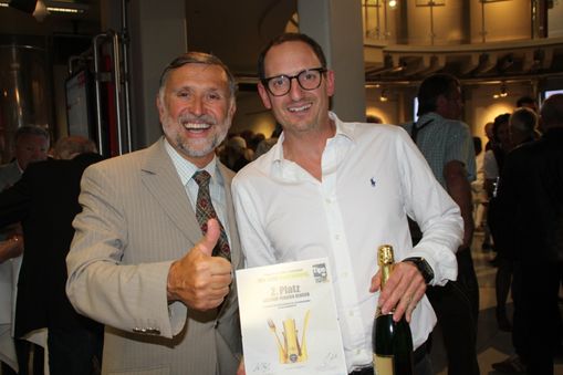 zwei Männer lachen gelassen in die Kamera der linke ist schon grau haarig und trägt Vollbart und streckt den Daumen hoch, der rechte ist mittleren Alters und trägt eine schwarz gerandete Brille und Schnauz- und drei Tage Bart und hält eine Flasche Sekt in der einen und den Award Lieblingswirt in Oberösterreich 2017 in der anderen