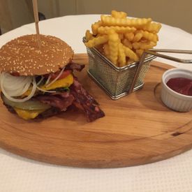 Servierplatte aus Holz auf weißem Tisch, darauf links der Rensch Burger mit Bacon, Gewürzgurke, Zwiebel, Tomate; daneben in einem Mini Frittier Sieb Pommes und daneben ein kleines weißes Schälchen mit Ketchup 