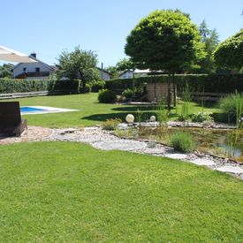 Blick in den gepflegten Garten des Gasthauses, links ist ein kleines Stück des Swimmingpools zu sehen und davor ein Sessel, rechts im Bild ist ein Teich zu sehen, dahinter Bäume