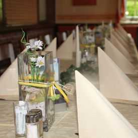 gedeckte Tage für eine Gesellschaft, dekorativer quadratisch-hoher Vase mit Sand gefüllt und weiß-gelben Blumen, beigefarbene Servietten und Kerzen, Tischdecke aus grünen Zweigen