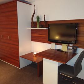 Blick auf den Kleiderschrank mit Sitzecke mit Möbeln in weiß und rotbrauner Holzoptik. Ein weißer Stuhl am Tisch über dem ein Flachbildfernseher hängt.Daneben ein weißes Regalbrett mit Pflanzen. Auf dem Tisch stehen Blumen in Dekovase