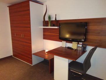 Blick auf den Kleiderschrank mit Sitzecke mit Möbeln in weiß und rotbrauner Holzoptik. Ein weißer Stuhl am Tisch über dem ein Flachbildfernseher hängt.Daneben ein weißes Regalbrett mit Pflanzen. Auf dem Tisch stehen Blumen in Dekovase
