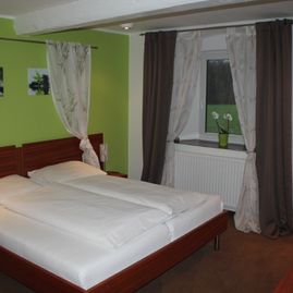 Doppelbett Zimmer mit lindgrüner Wand hinter dem Bett un Dekovorhang an der Wand der das Bett einrahmt. Neben dem Bett ist die Fensterwand mit einzelnem Fenster eingerahmten von weißen Gardinen und grauem Vorhang. Auf dem Fensterbrett steht eine Vase mit Blumen.