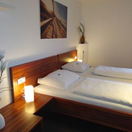 Blick auf das Bett in roter Holzoptik mit weißem Bettbezügen, warmen Nachttischlicht und einem Bild an der Wand über dem Bett.