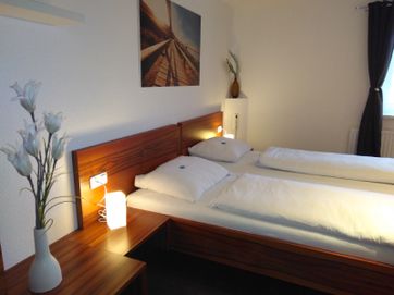 Blick auf das Bett in roter Holzoptik mit weißem Bettbezügen, warmen Nachttischlicht und einem Bild an der Wand über dem Bett.